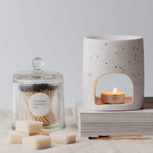White ceramic wax melt burner with a lit candle inside, sitting on a pile of magazines, with a branded cloche of matches, and scattered wax melt cubes
