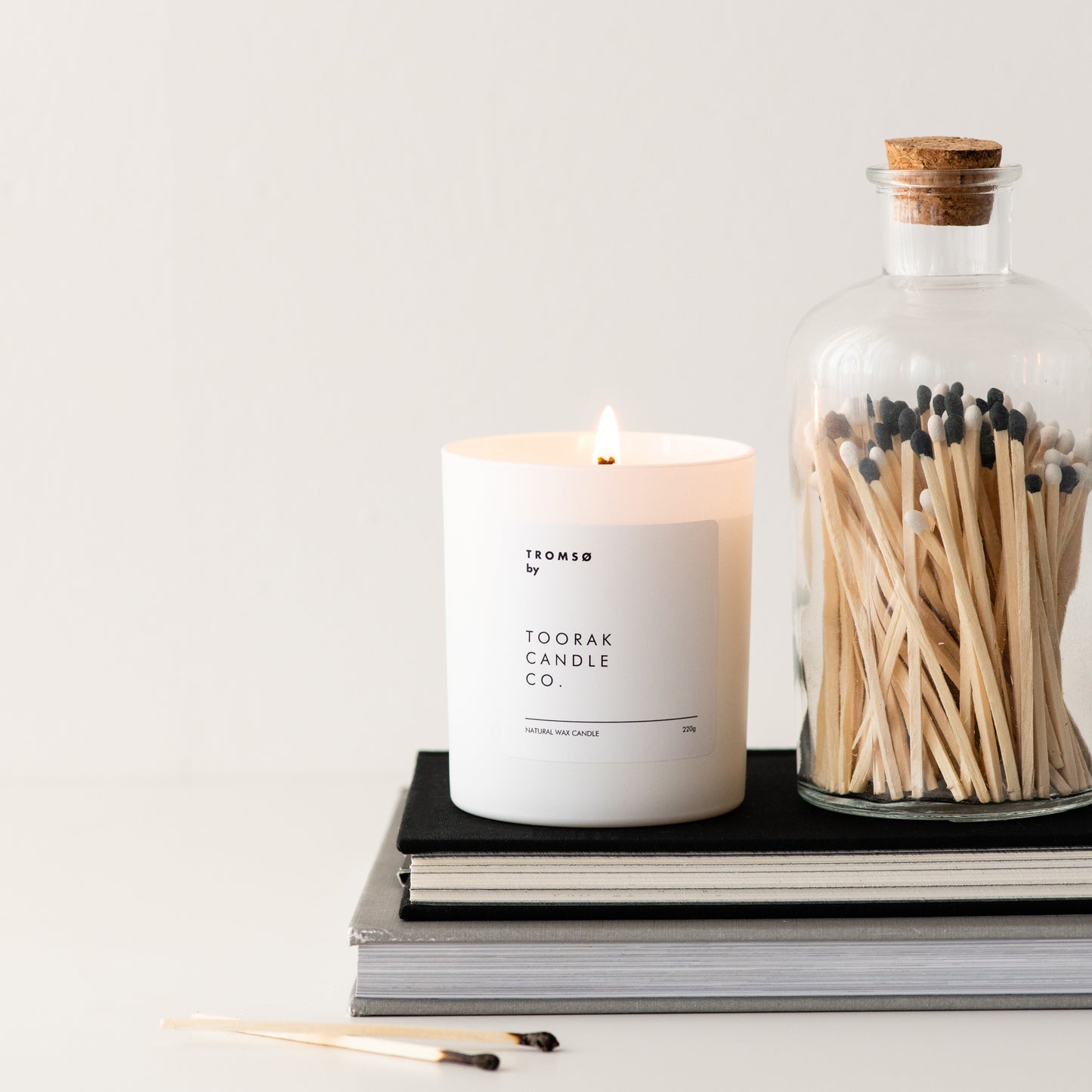 Lit TROMSO natural wax candle on a tack of books, next to a jar of black and white-tipped wooden matches with a cork top