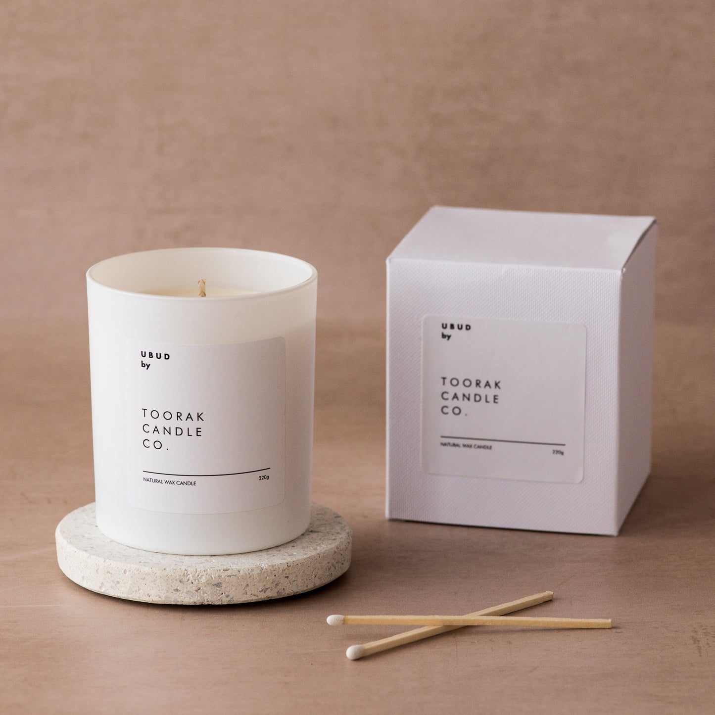 UBUD natural wax candle on a ceramic coaster, next to an UBUD box, with two white-tipped matches, on a light brown background