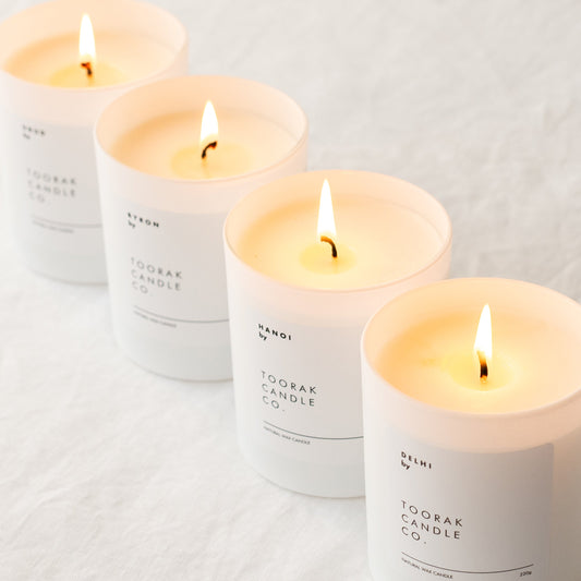 Four lit luxury natural wax candles in a diagonal layout on a crinkled linen white background