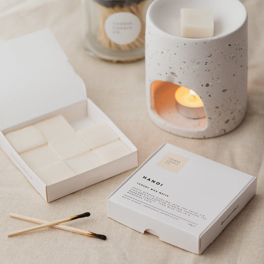 HANOI wax melts with wax melt burner, next to an open box of wax melts and some matches, on a cream linen background