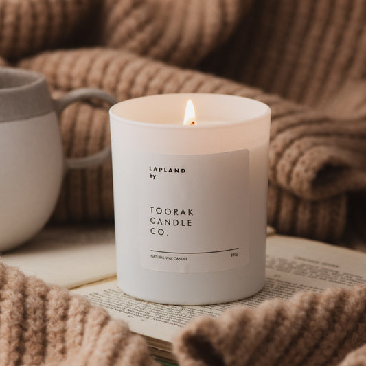 Cosy image of LAPLAND Natural Wax Candle lit, sitting on an open book, surrounded by a cream knitted blanket and a mug