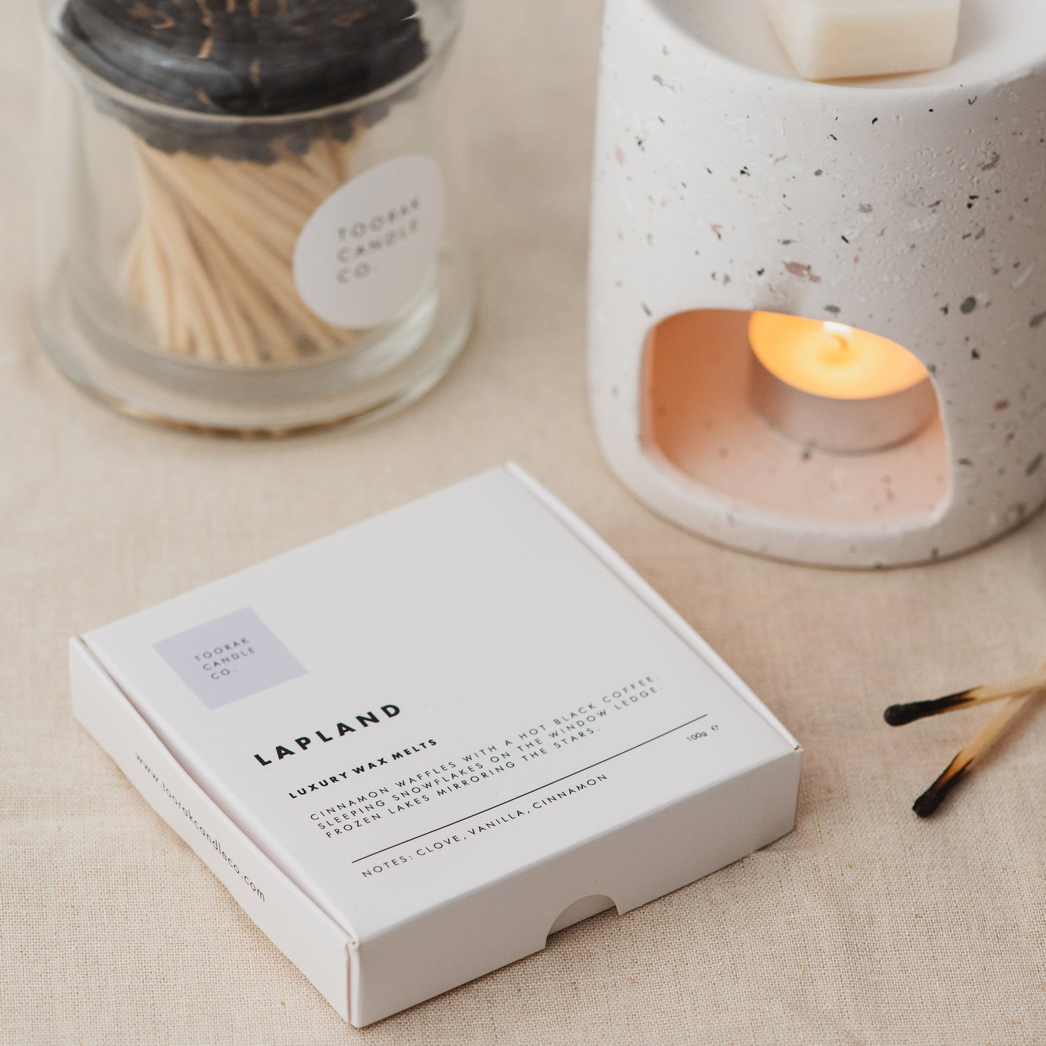 LAPLAND wax melts on a cream linen background with a lit wax melt burner and branded matches 