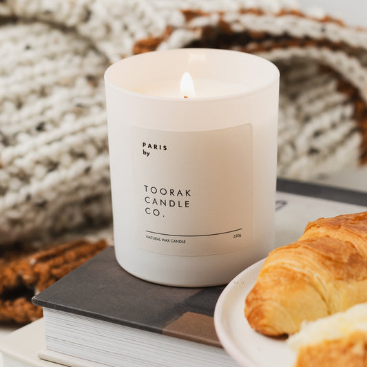 White scented candle with 'Paris by Toorak Candle Co.' label on a surface with books and pastries.