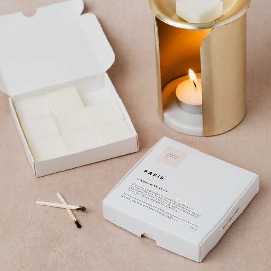 Paris croissant-scented wax melts in a box with matches and a lit candle in a gold wax melt burner on a beige surface.