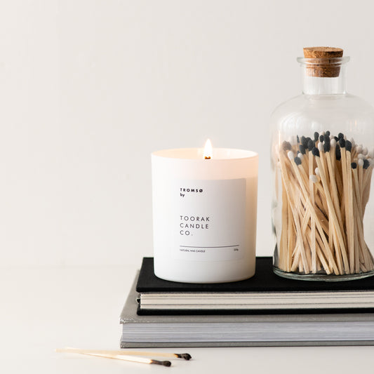 Lit TROMSO natural wax candle on a tack of books, next to a jar of black and white-tipped wooden matches with a cork top
