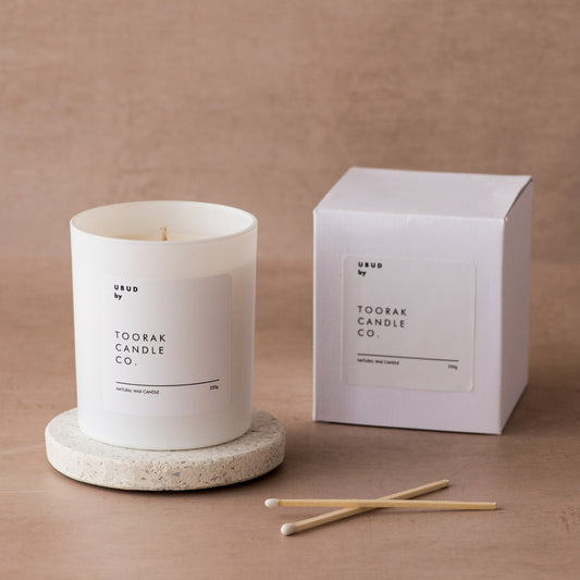 UBUD natural wax candle on a ceramic coaster, next to an UBUD box, with two white-tipped matches, on a light brown background 